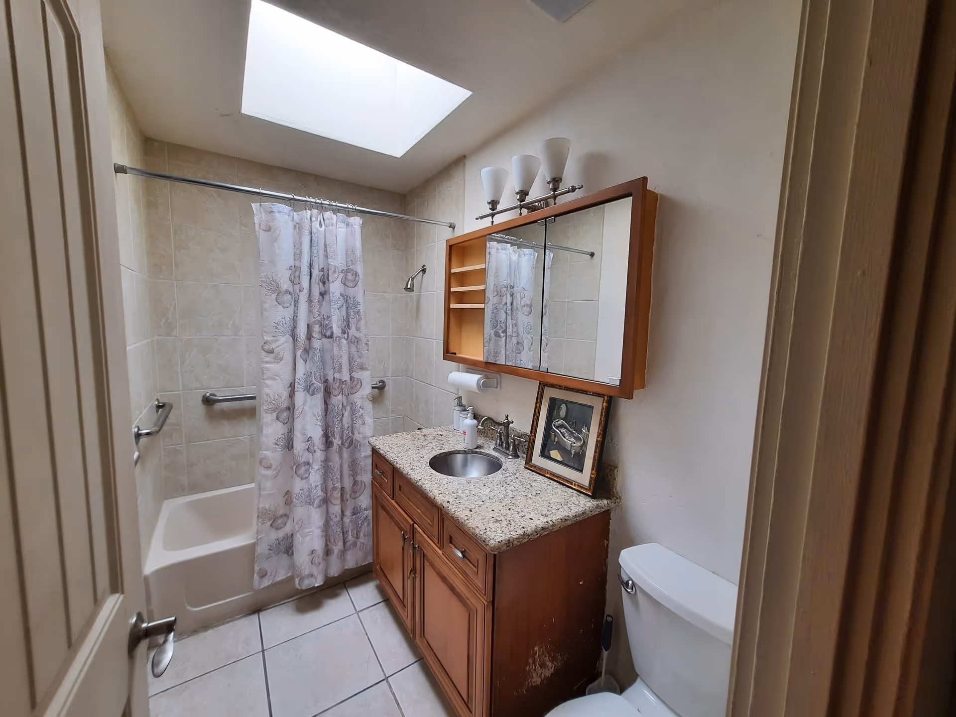 A small bathroom with a bathtub and shower combination on the left, featuring a shower curtain with seashell and coral patterns. The walls around the tub are tiled. There is a wooden vanity with a granite countertop and a round sink in the center. Above the vanity is a large mirror with a wooden frame and three light fixtures on top. A framed picture is leaning against the wall on the countertop. To the right is a white toilet with a toilet brush beside it. The floor is tiled, and there is a skylight above providing natural light.