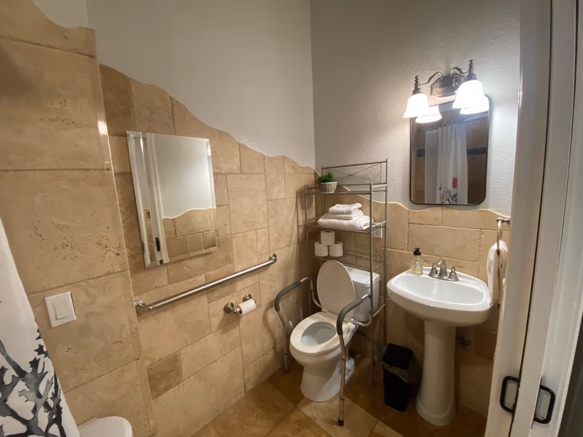 A small bathroom with beige tiled walls and floor. There is a white toilet with safety rails on both sides, a metal shelving unit above the toilet holding towels and toilet paper, a white pedestal sink with a soap dispenser, a mirror with two light fixtures above it, and a towel ring with a white towel. A grab bar is mounted on the wall next to the toilet, and a shower curtain is partially visible on the left side.