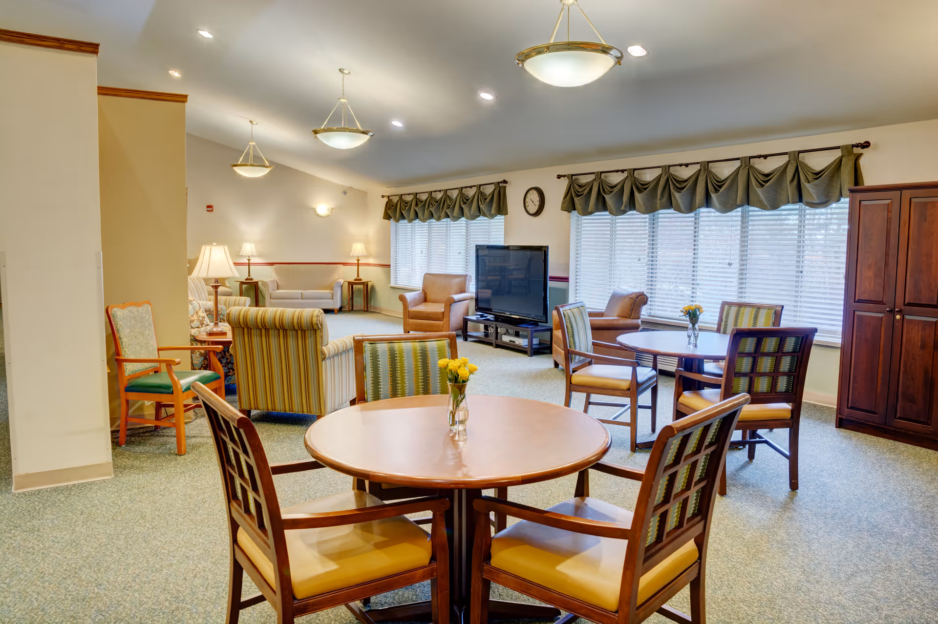 Communal lounge with round tables and chairs, sofas, armchairs, a TV and large windows with valances.