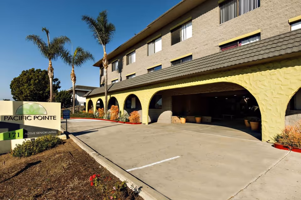 Exterior view of the Pacific Pointe senior living facility showing a three-story building with arched yellow accents on the ground floor, palm trees, and a driveway leading to the entrance. A sign in front reads 'Pacific Pointe 171 Active Senior Living.'
