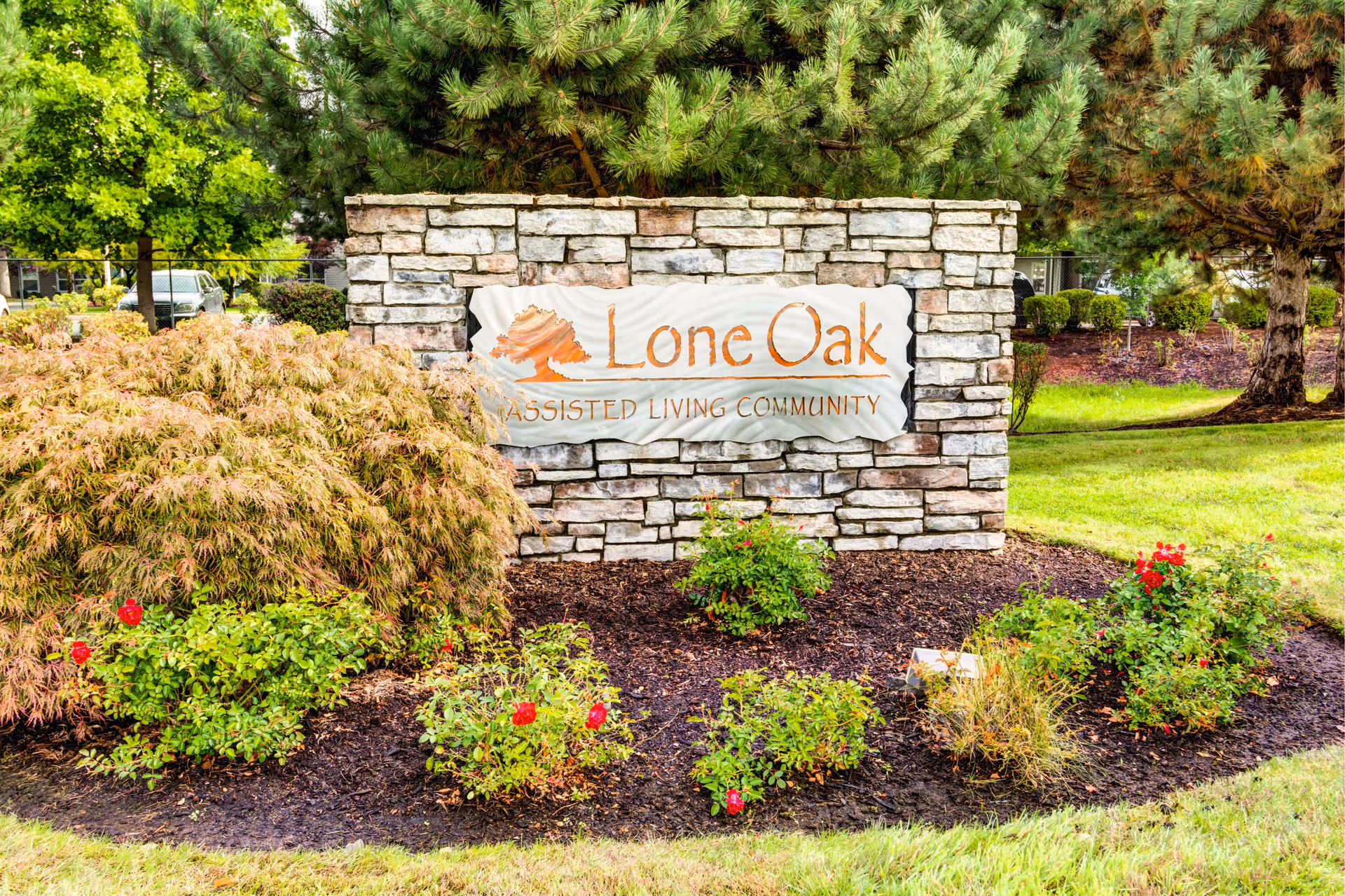 Stone sign for Lone Oak Assisted Living Community surrounded by green grass, bushes, and trees in a landscaped outdoor area.