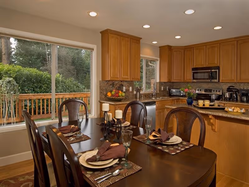 Dining table set for four in an open-plan kitchen with wooden cabinets and large windows overlooking a deck and trees.