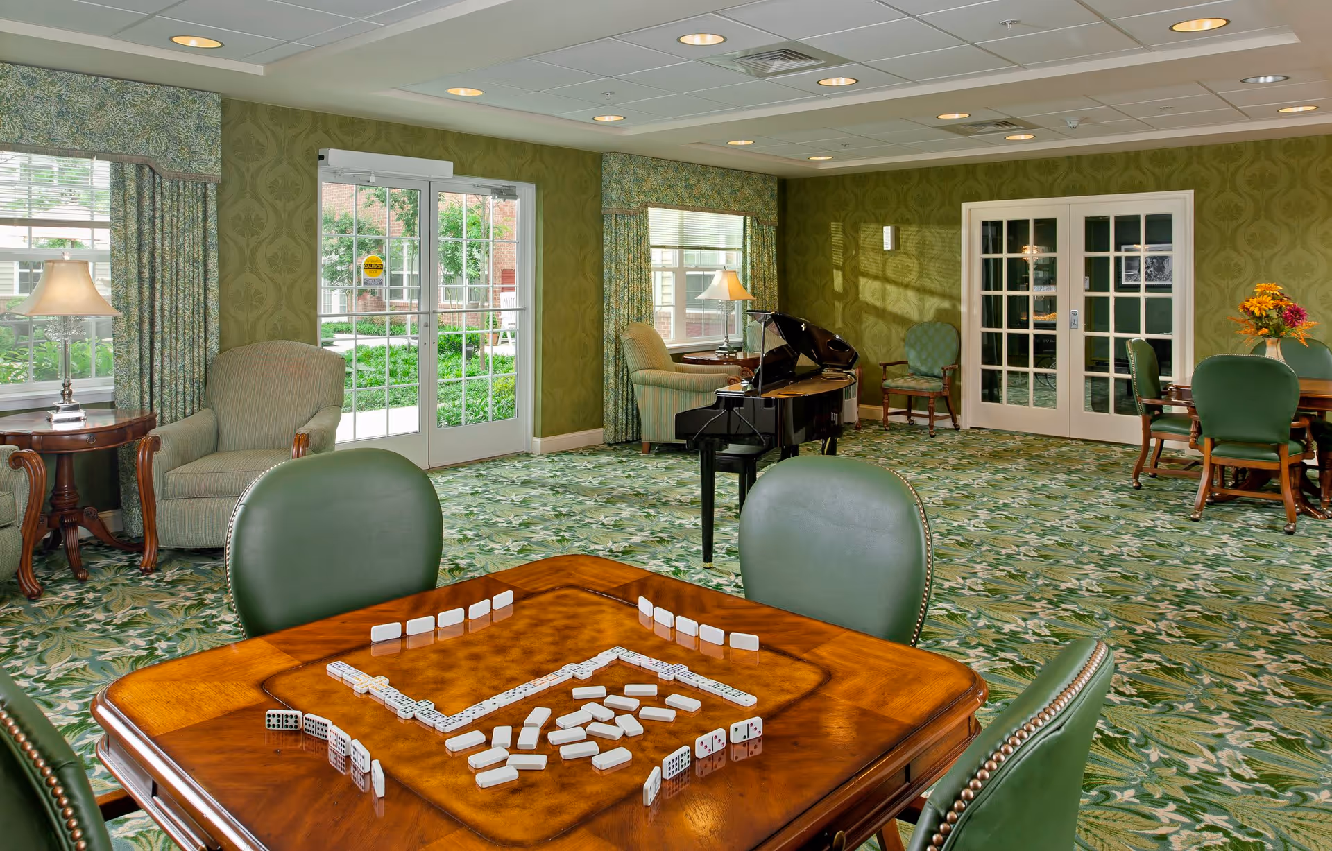 A spacious common room with green patterned carpet and wallpaper, featuring a wooden table with dominoes set up for a game, green cushioned chairs, a grand piano, armchairs, side tables with lamps, and large windows and glass doors letting in natural light.