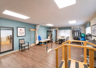 Physical therapy room with parallel bars, wooden practice stairs, a treatment table, exercise equipment, therapy balls and seating.