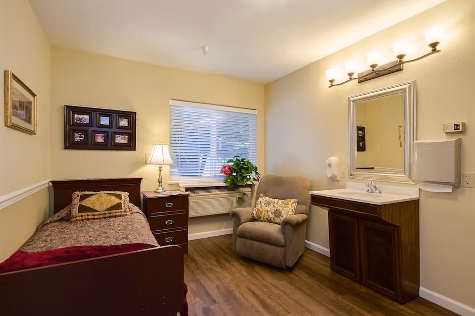 Cozy single-occupancy bedroom with a bed, nightstand, armchair, window, and a sink with mirror.