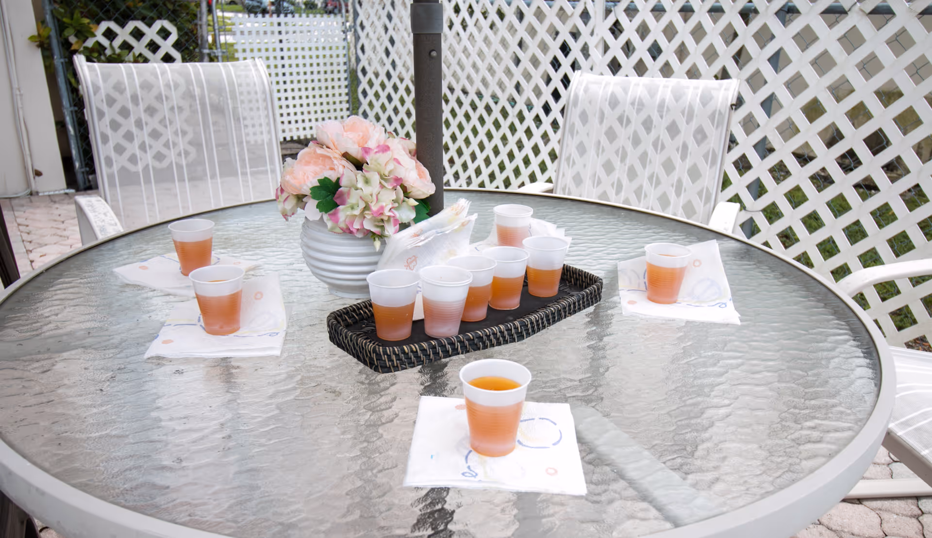 Outdoor round glass table with several plastic cups filled with a light brown beverage placed on napkins, a white vase with pink and white flowers in the center, surrounded by white lattice fencing and white patio chairs.