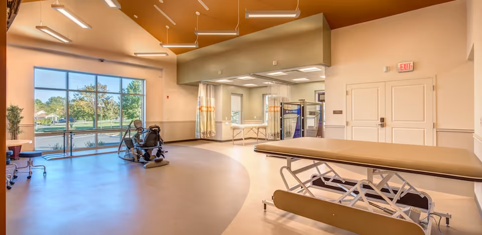 A spacious rehabilitation room with a large window showing outdoor greenery. The room contains a therapy exercise bike, an adjustable treatment table, and a curtained area with additional tables and equipment. The ceiling has modern lighting fixtures and the walls are painted in neutral tones.
