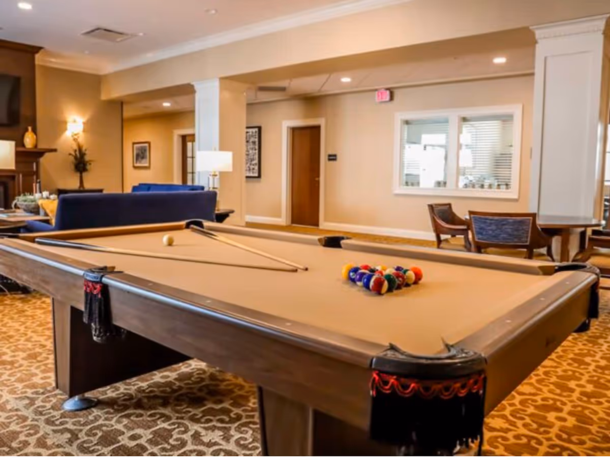 A well-lit senior living facility common area featuring a pool table with billiard balls and cues arranged on it. In the background, there are blue sofas, a fireplace, lamps, chairs, and a window looking into another room. The room has beige walls and patterned carpet flooring.