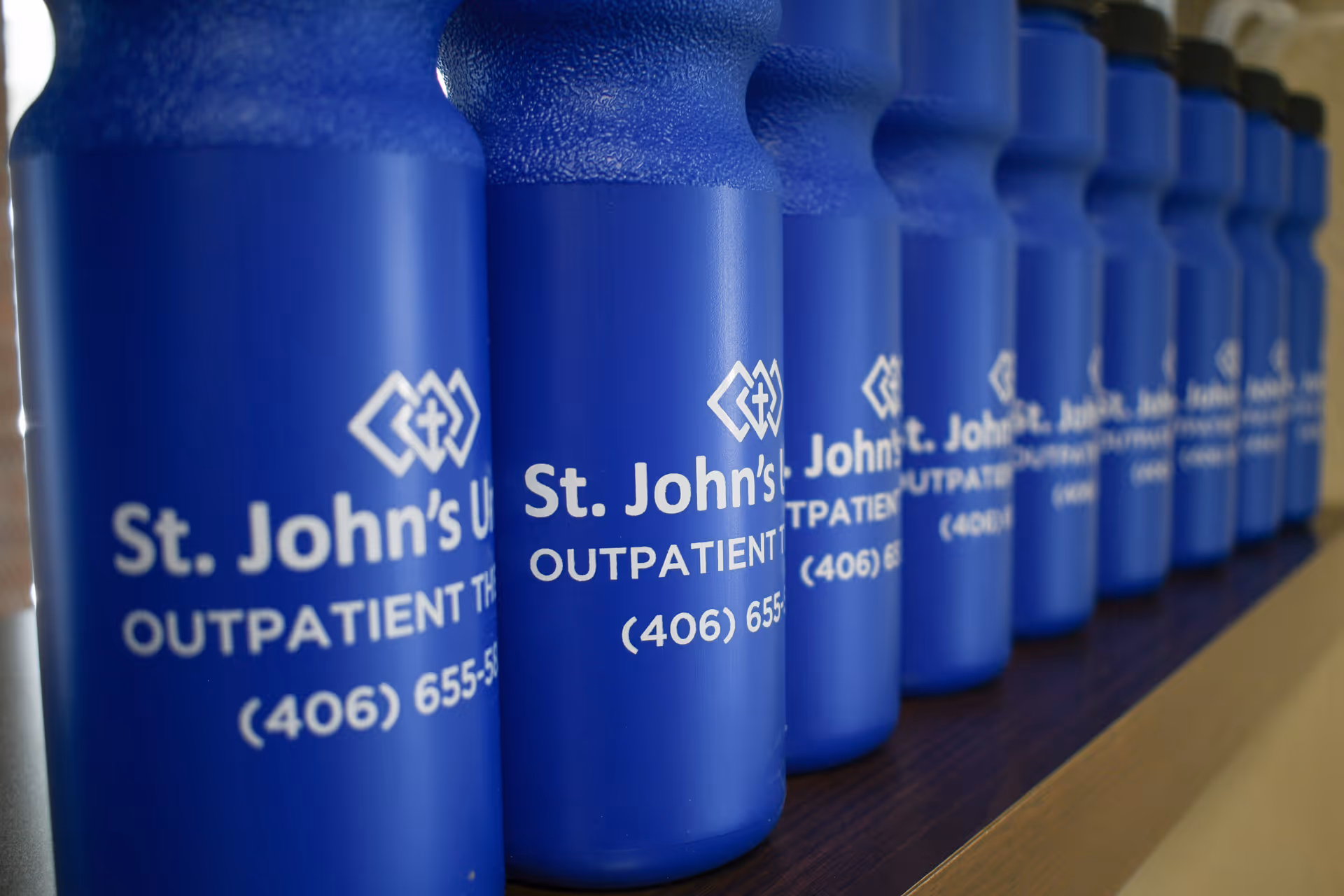 A row of blue water bottles lined up on a shelf, each labeled with 'St. John's United Outpatient Therapy' and a phone number.