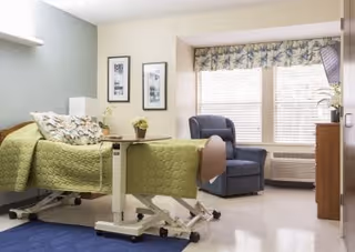 Well-lit nursing facility room with an adjustable bed, armchair by a large window, dresser, and bedside table.