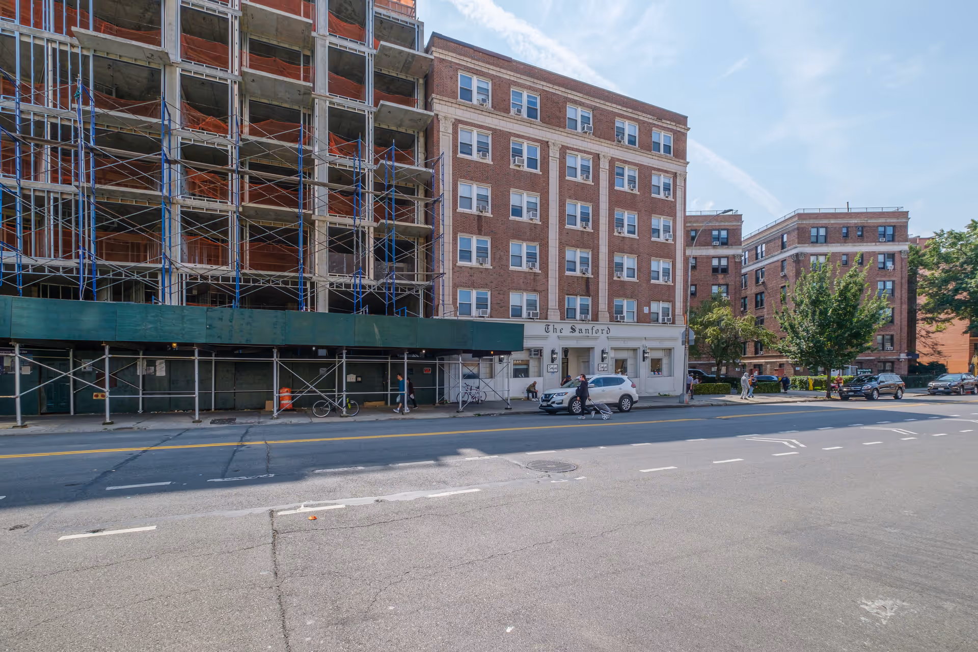 Street view of a multi-story brick building named The Sanford with adjacent construction scaffolding and several people walking on the sidewalk. Cars are parked along the street and trees are visible in the background under a partly cloudy sky.
