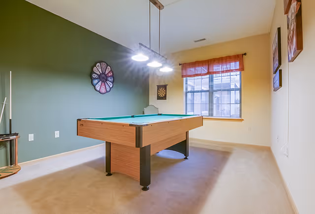 A well-lit game room with a pool table in the center, a green accent wall with a decorative clock, a dartboard on the wall, and a window with an orange valance letting in natural light.
