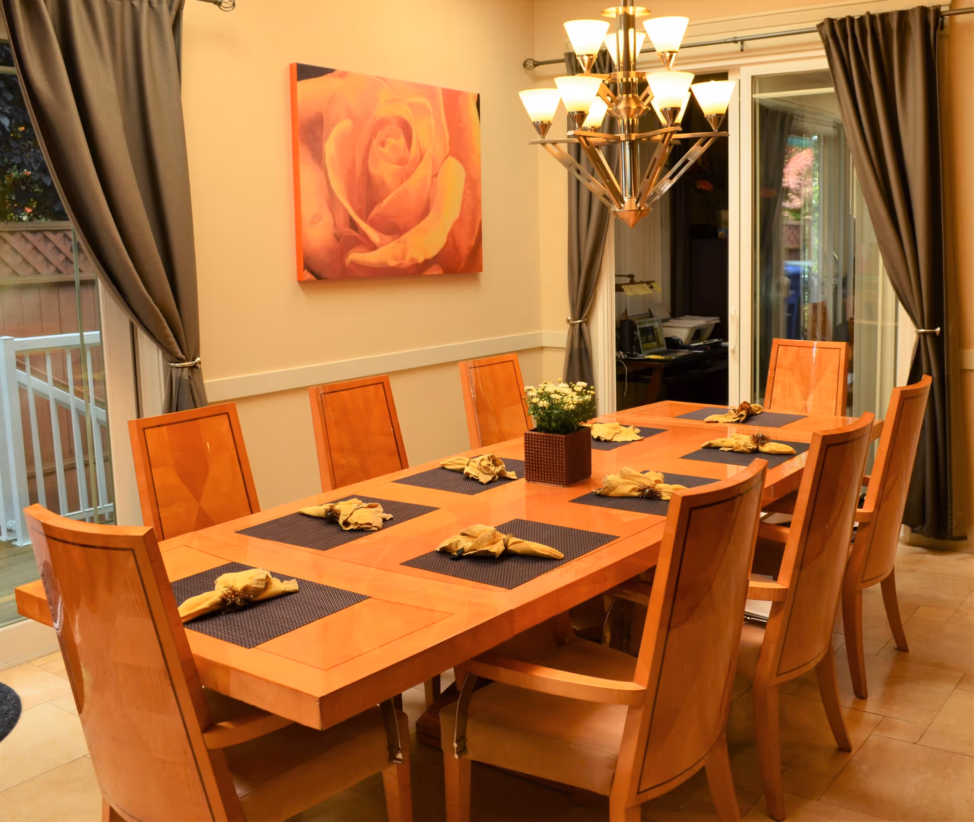 A dining room with a large wooden table set with eight chairs. Each place setting has a dark placemat and a folded yellow napkin. A small potted plant with white flowers is centered on the table. The room has beige walls, a large painting of a rose, and a modern chandelier with multiple lights. There are large windows with gray curtains on two sides, one showing an outdoor deck area.