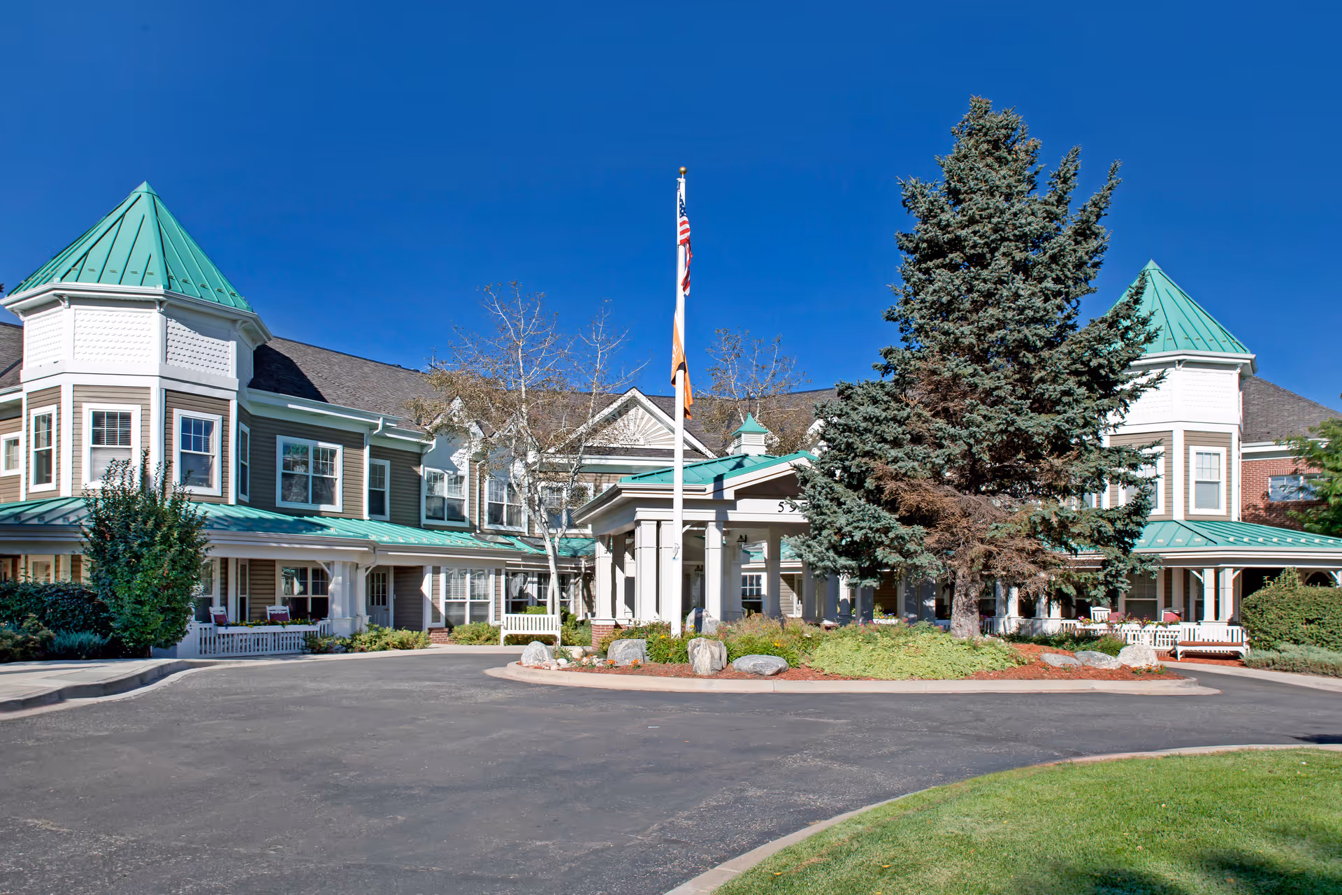 Front exterior of a senior living facility with green-roofed turrets, a flagpole, circular driveway and landscaped entrance.