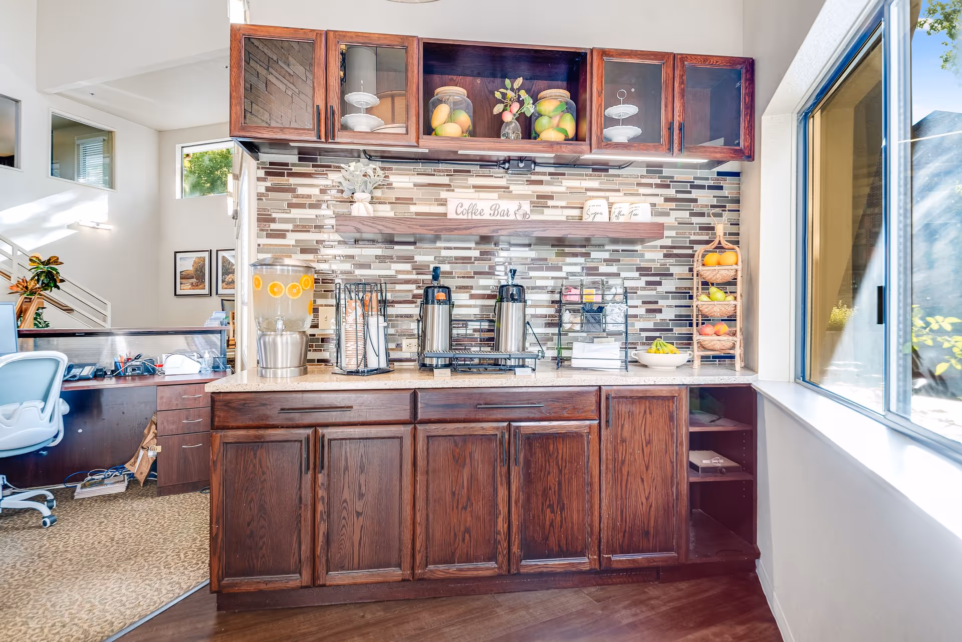 Small wooden coffee bar with stainless beverage dispensers, glass jars of fruit, and cabinetry against a tiled backsplash next to a window.