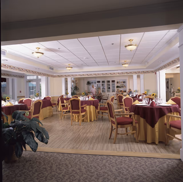 A spacious dining room in a senior living facility with round tables covered in yellow and burgundy tablecloths, set with glassware and napkins. Several chairs with wooden frames and burgundy cushions surround the tables. The room has a light wood floor, decorative ceiling lights, and large windows letting in natural light. A few elderly residents and a staff member are visible in the background near a cabinet with dishes and plants.