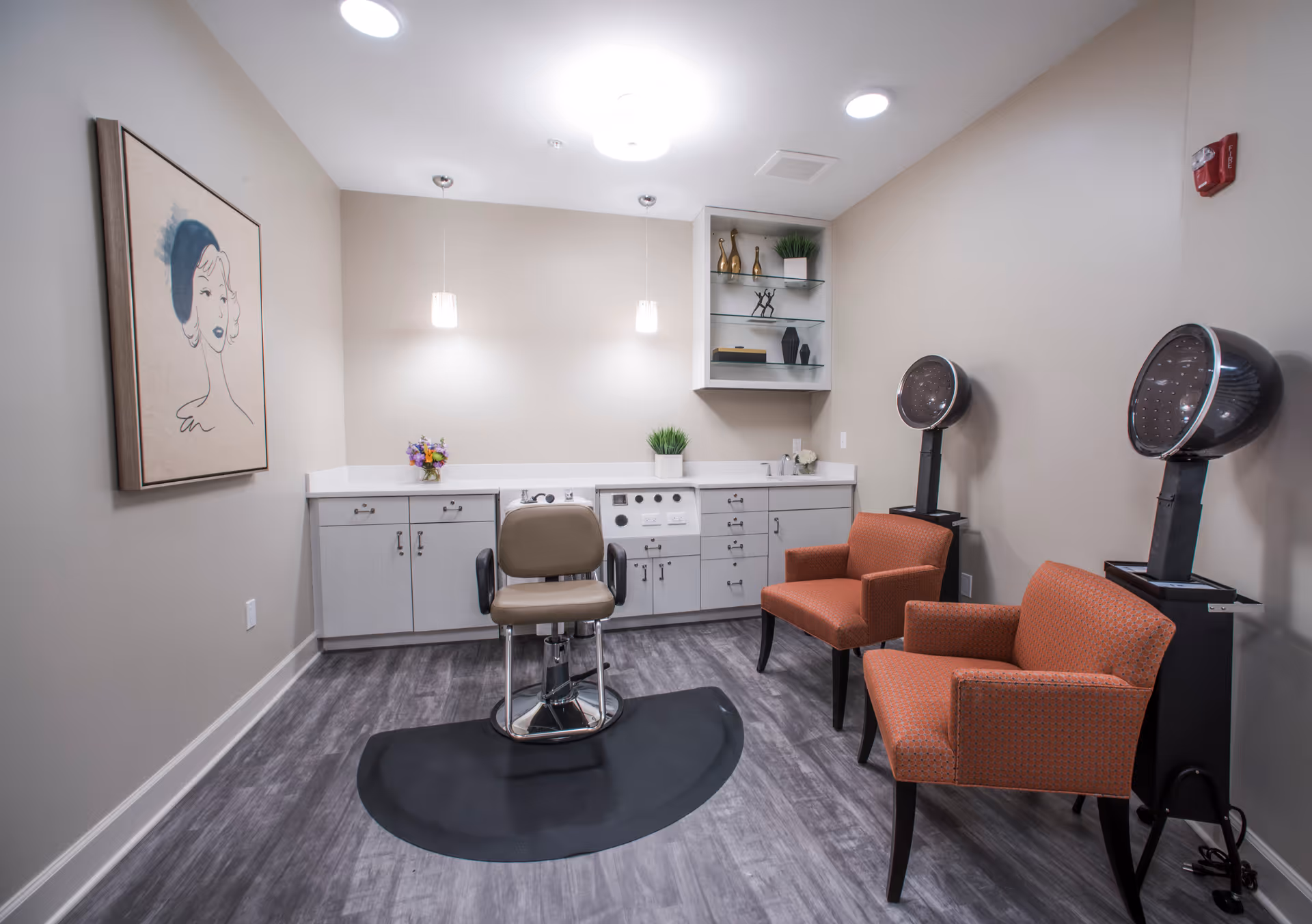 A small salon room with a beige salon chair on a black mat in the center, two orange upholstered chairs on the right, and two hair dryer stations behind the chairs. The back wall has white cabinets, a countertop with a small flower arrangement, and shelves with decorative items. A framed artwork of a woman's face hangs on the left wall.