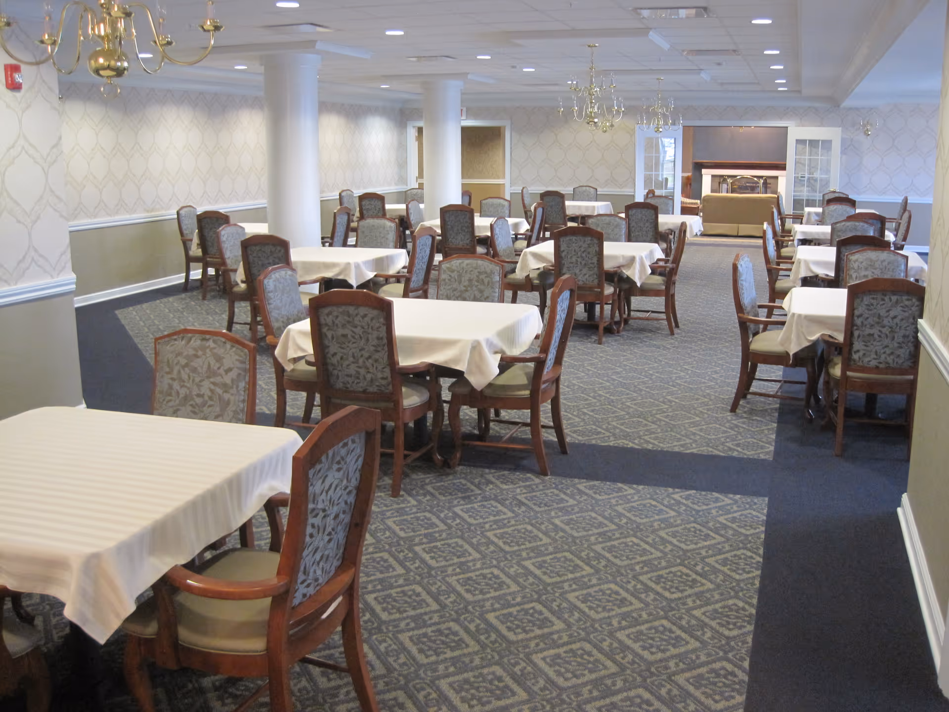 A spacious dining room with multiple tables covered in white tablecloths, each surrounded by wooden chairs with patterned upholstery. The room features carpeted floors with a geometric design, beige patterned wallpaper, white pillars, and brass chandeliers hanging from the ceiling. In the background, there is a seating area with a fireplace and a sofa.