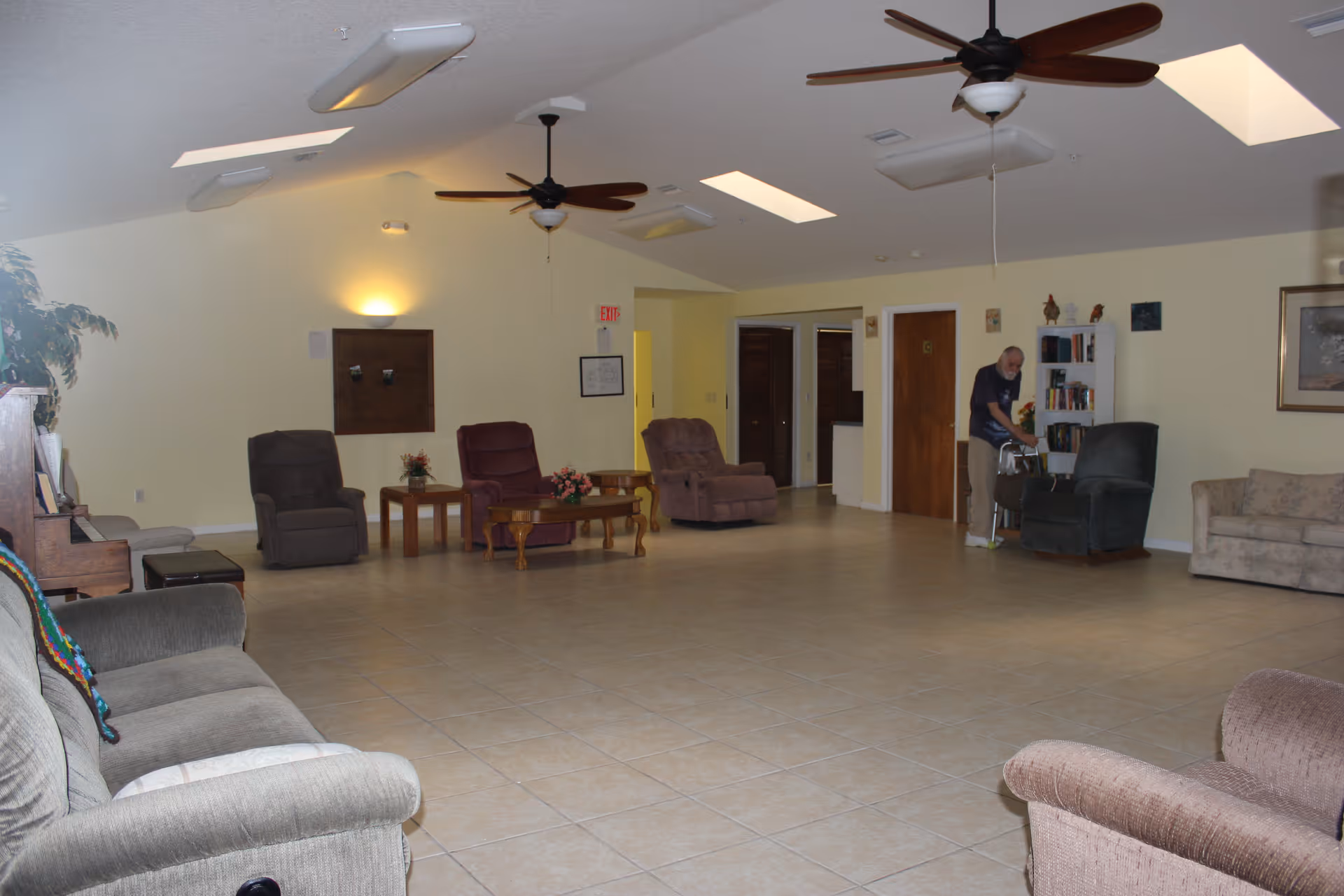 A spacious living room area with tiled floors, several armchairs and sofas arranged around wooden coffee tables, ceiling fans, skylights, and a person cleaning near a bookshelf.