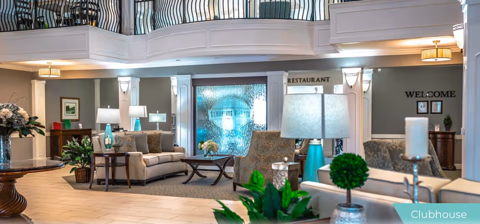A spacious and well-lit senior living facility clubhouse with comfortable seating including sofas and armchairs, decorative plants, table lamps, and a water feature with the facility's name. The area has a welcoming ambiance with a sign indicating the restaurant and a 'WELCOME' sign on the wall.