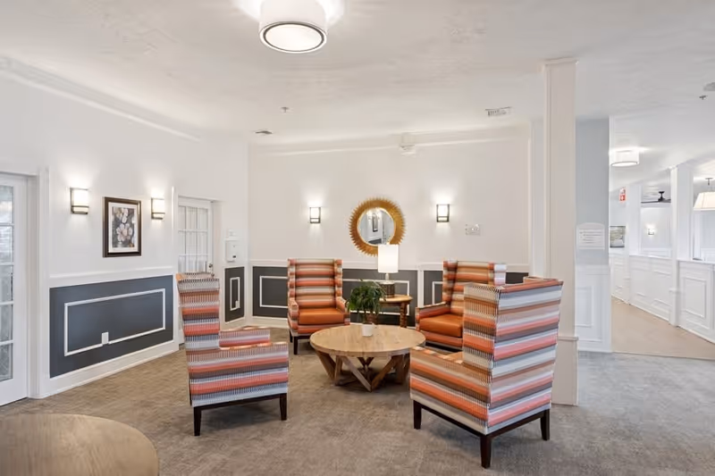 A cozy seating area in a senior living facility with four striped armchairs arranged around a round wooden coffee table. The room features white walls with dark gray wainscoting, a round decorative mirror, wall sconces, and a small plant on the table. The space is well-lit with ceiling lights and has a carpeted floor.
