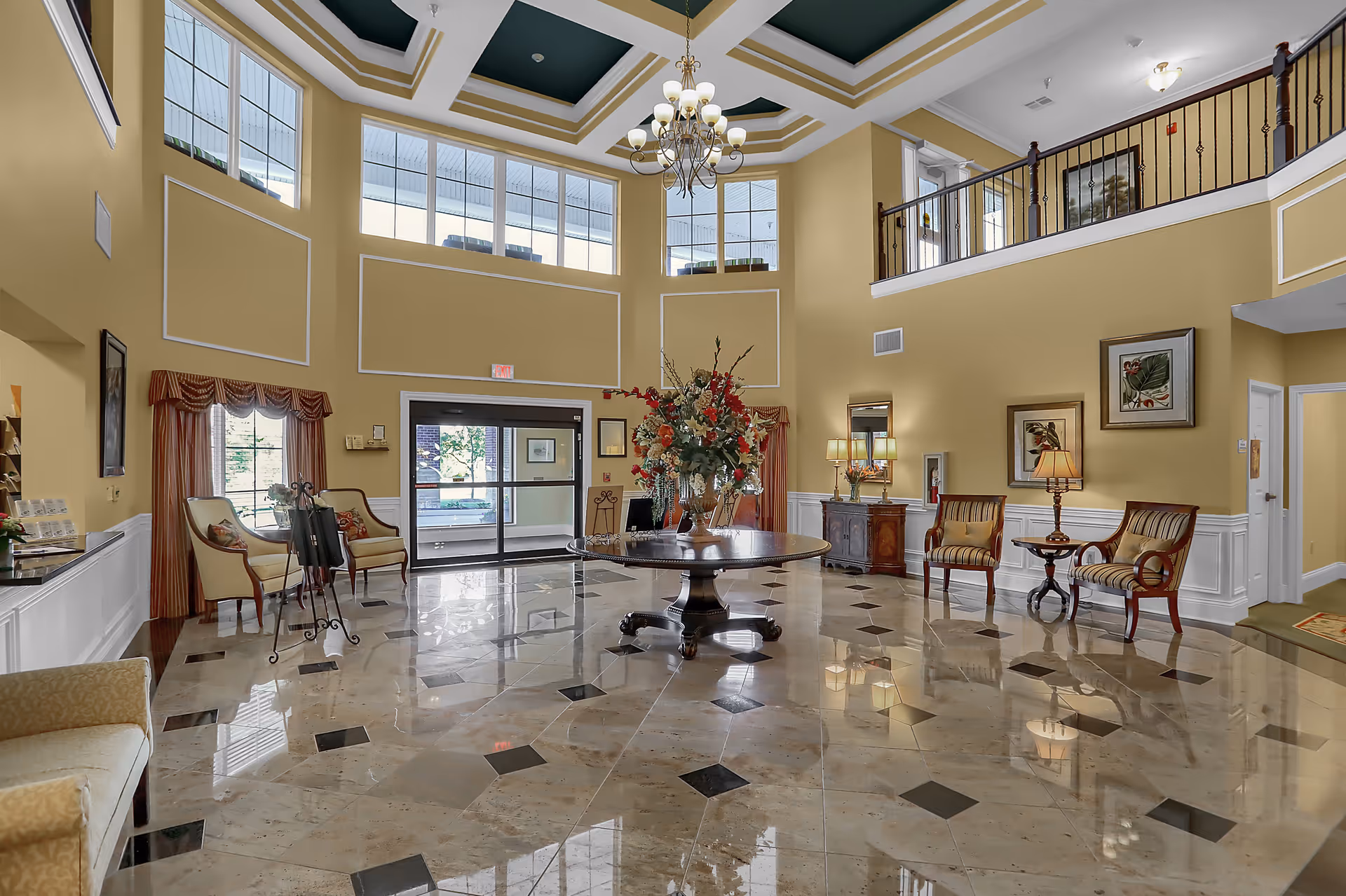 Spacious and elegant lobby area with high ceilings, large windows, and a chandelier. The floor is polished with a patterned tile design. There is a round table with a large floral arrangement in the center. Several chairs and small tables with lamps are arranged along the walls. The walls are painted yellow with white wainscoting and decorated with framed artwork.