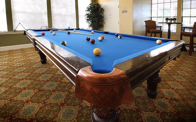 A blue-felt pool table in a carpeted common room with windows, chairs, and a plant.