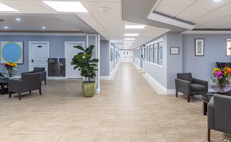 Wide, well-lit interior hallway with seating areas, potted plants, and doors lining the corridor in a senior living facility.