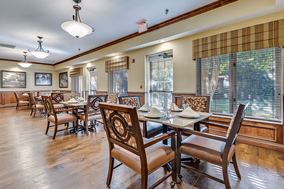 Well-lit dining room with wooden tables and upholstered chairs set with napkins and glassware and large windows along one wall.