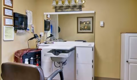 Interior view of a hair salon station with a large mirror, a white countertop with a built-in sink, a black salon chair, hair care products, and a wall-mounted hair dryer. The walls are painted beige and decorated with framed certificates and a floral picture.