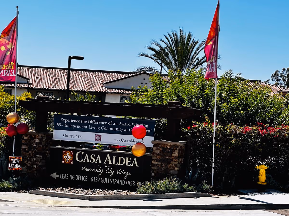 Entrance sign for Casa Aldea at University City Village with balloons, flags, and the building roof behind it.