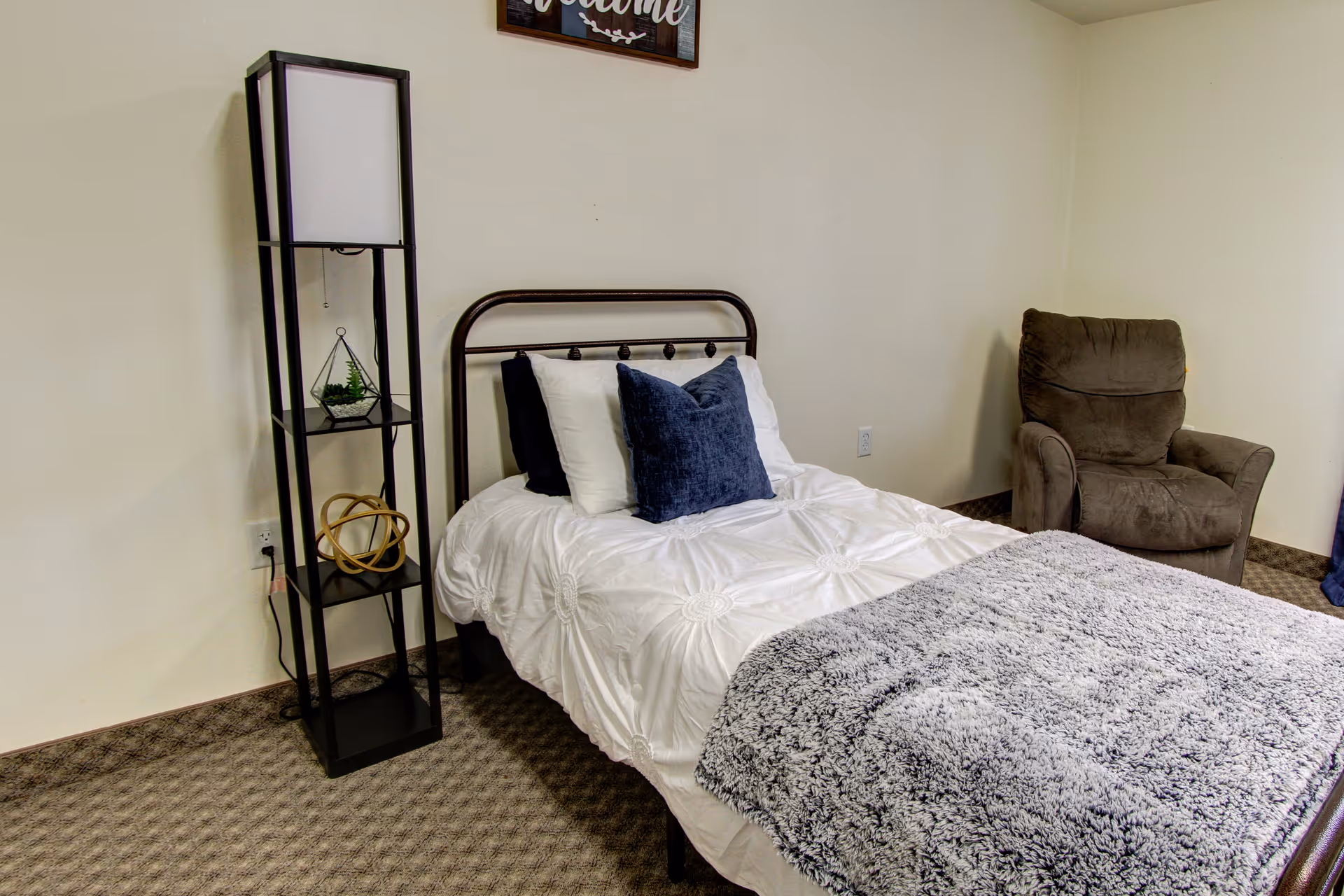 A simple bedroom with a single bed dressed in white bedding and blue pillows, a tall black shelving lamp, and a brown recliner chair.