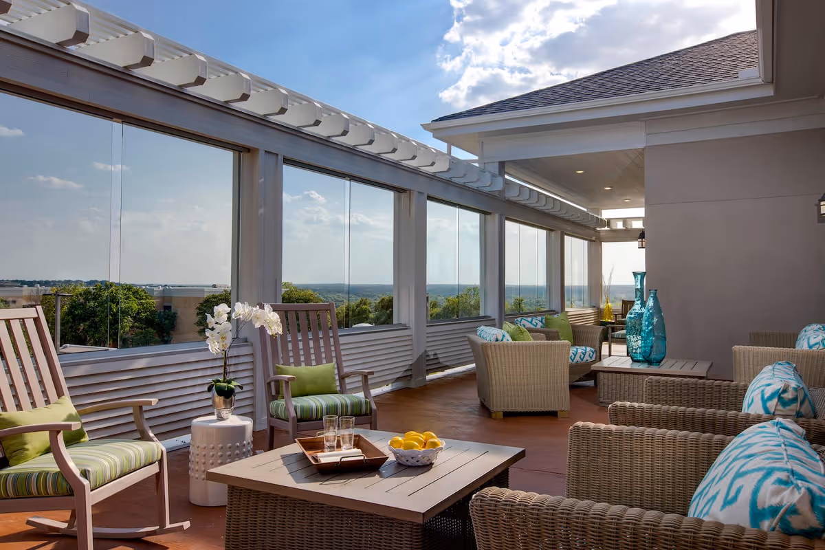 A spacious outdoor patio area with wicker chairs and sofas adorned with blue and green patterned cushions. There are two wooden rocking chairs with green striped cushions, a small white side table with a white orchid plant, and a coffee table with a tray holding glasses and a bowl of lemons. The patio is enclosed with large glass panels offering a scenic view of trees and a distant horizon under a partly cloudy sky.