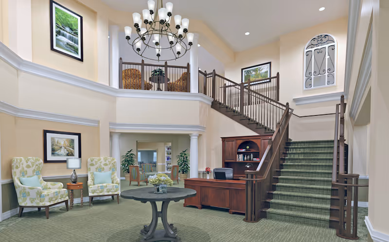 Spacious two-story reception lobby with a central round table, upholstered chairs, a wooden reception desk, and a staircase leading to an upper balcony.