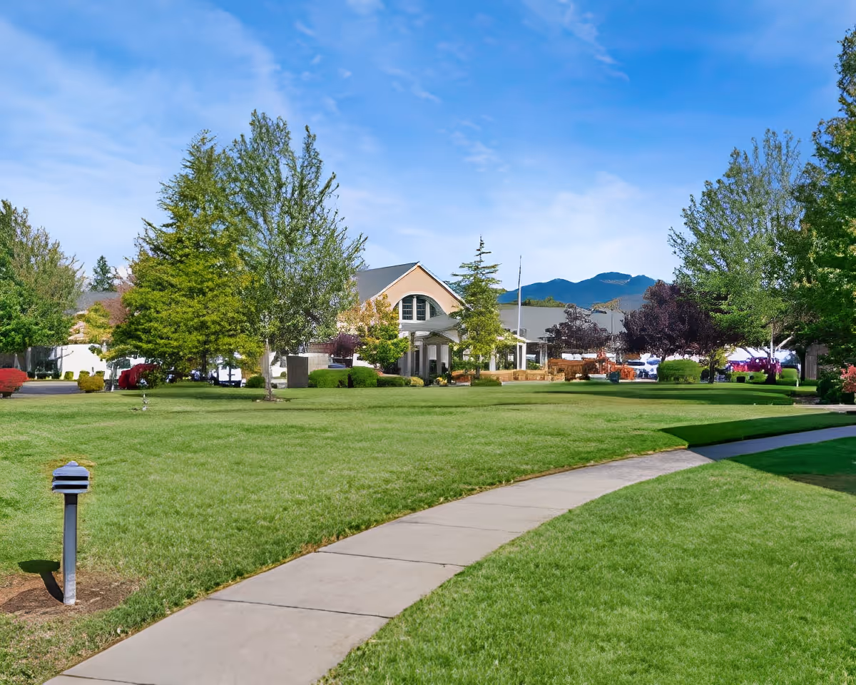 A well-maintained outdoor area at Cascades of Grants Pass featuring a wide green lawn, a curved concrete walkway, various trees, and a building with a covered entrance in the background under a blue sky with some clouds.