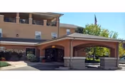 Exterior view of a multi-story assisted living facility with a covered entrance and an American flag flying on a flagpole. The building has beige walls and several windows, with trees and landscaping around the entrance.