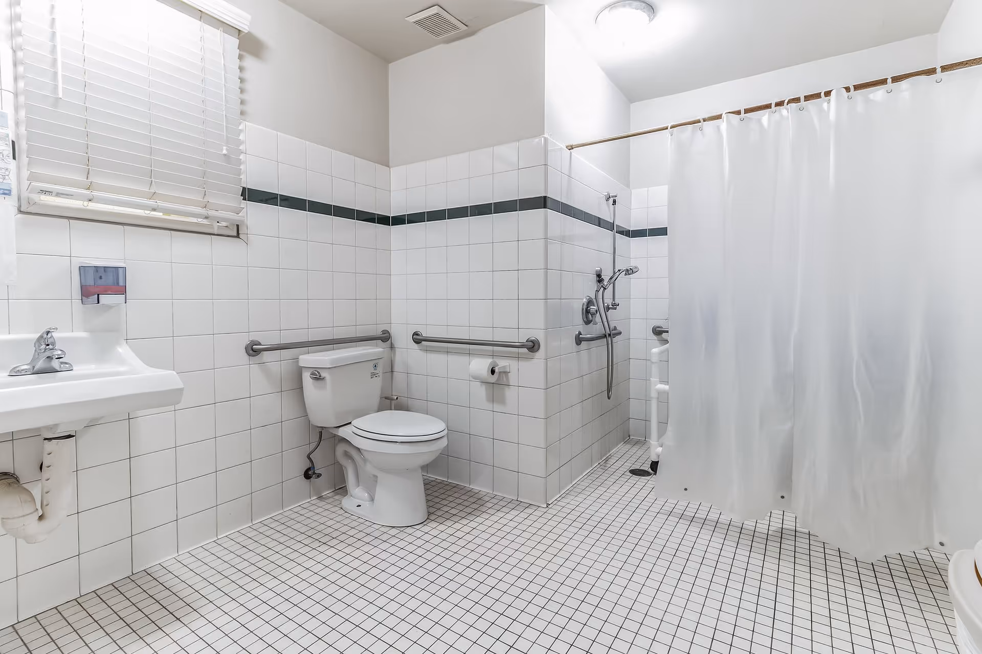 A clean, accessible bathroom with white tiled walls and floor. The bathroom features a white toilet with grab bars on both sides, a white sink with a faucet, a window with white blinds, and a shower area with a white curtain and a handheld showerhead.