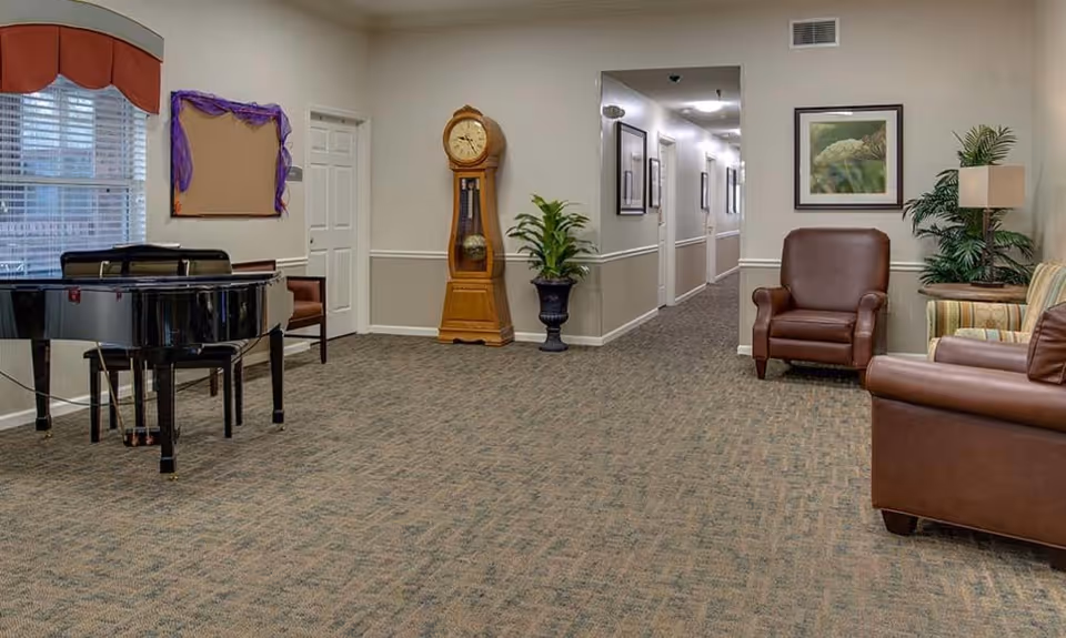 A spacious common area in a senior living facility featuring a black grand piano on the left, a tall wooden grandfather clock, a potted plant, and a hallway leading to other rooms. On the right side, there are brown leather armchairs, a striped sofa, a side table with a lamp, and framed artwork on the wall.