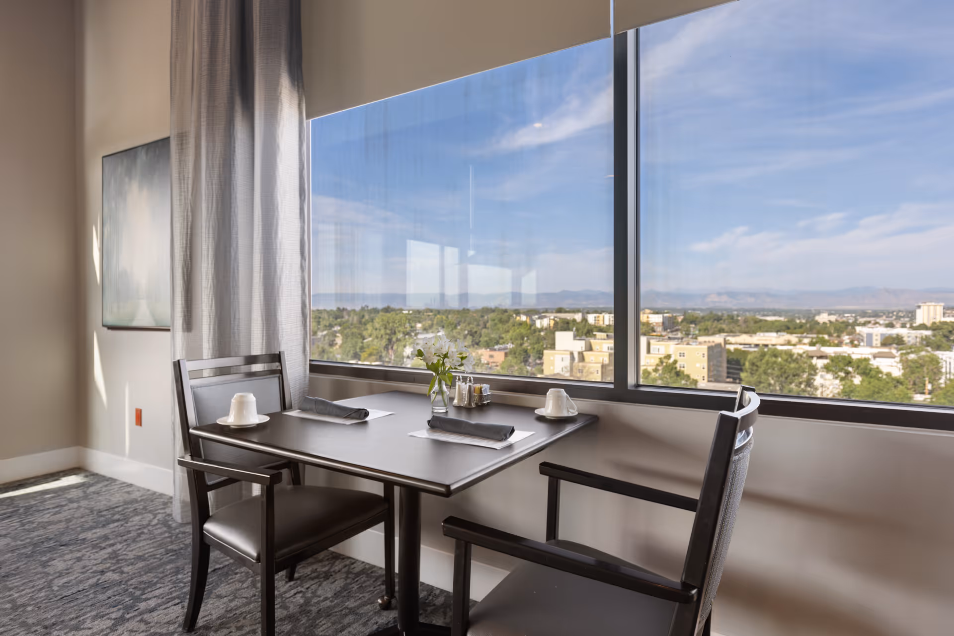 A small dining table set for two with black chairs, white cups, napkins, and a small vase with flowers, positioned next to a large window offering a panoramic view of a cityscape and distant mountains under a blue sky.
