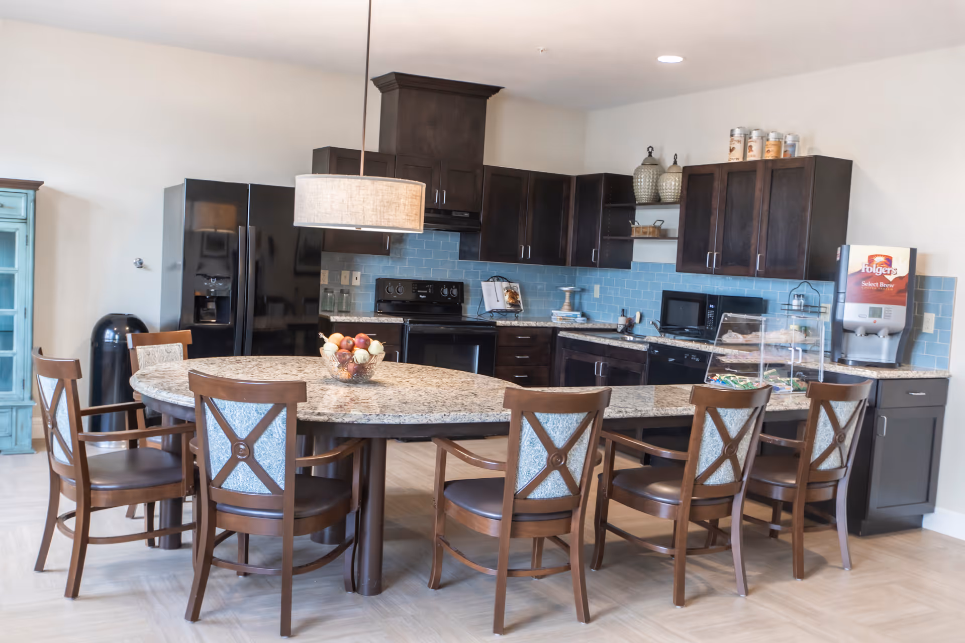 A modern kitchen area with dark wood cabinets, a black refrigerator, stove, microwave, and dishwasher. A large granite countertop island with six wooden chairs surrounds the island. A bowl of fruit is on the island, and a coffee machine is on the right side countertop. The backsplash is light blue tile, and a hanging light fixture is above the island.