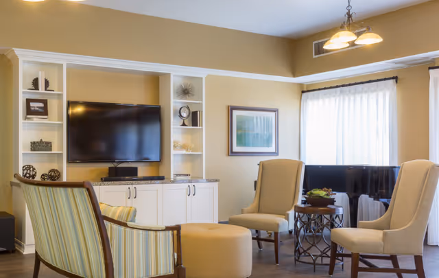 A cozy living room area with a flat-screen TV mounted on a white built-in shelving unit. The room features a striped armchair, two beige upholstered chairs, a round ottoman, and a small round table with a plant on it. Large windows with sheer white curtains allow natural light to fill the space. A framed picture hangs on the wall, and a chandelier with three lights is mounted on the ceiling.