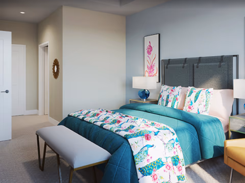 A bright and cozy bedroom with a neatly made bed featuring a teal quilt and floral peacock-themed bedding. The room has a padded headboard, two bedside tables with lamps, a bench at the foot of the bed, and a framed floral artwork on the wall. The walls are painted in light neutral tones, and there is a door leading to another room or hallway.