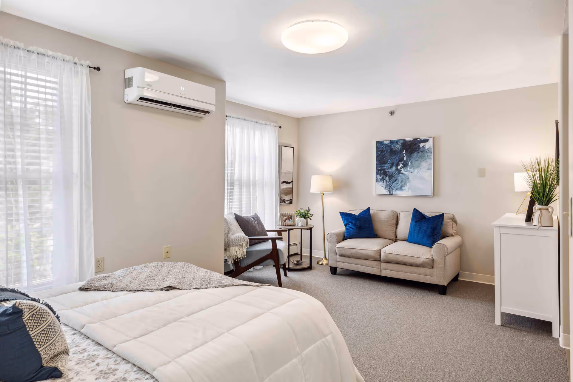 A bright and cozy senior living bedroom featuring a neatly made bed with white and patterned bedding, a beige loveseat with two blue pillows, a wooden armchair with cushions, a floor lamp, a small side table with a plant and picture frame, a white dresser with a lamp and plant, and two large windows with sheer white curtains allowing natural light to fill the room.