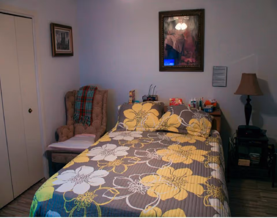 Bedroom with a bed dressed in a yellow-and-gray floral comforter, an armchair with a plaid throw, bedside tables with a lamp and personal items, and framed artwork on the wall.