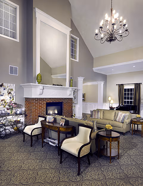 A spacious and elegantly decorated senior living facility common area featuring a brick fireplace with a large white mantel and mirror above it. The room has high ceilings with a decorative chandelier, comfortable seating including a sofa and armchairs, side tables with flowers, and a patterned carpet. There are windows with dark curtains and soft lighting creating a warm and inviting atmosphere.
