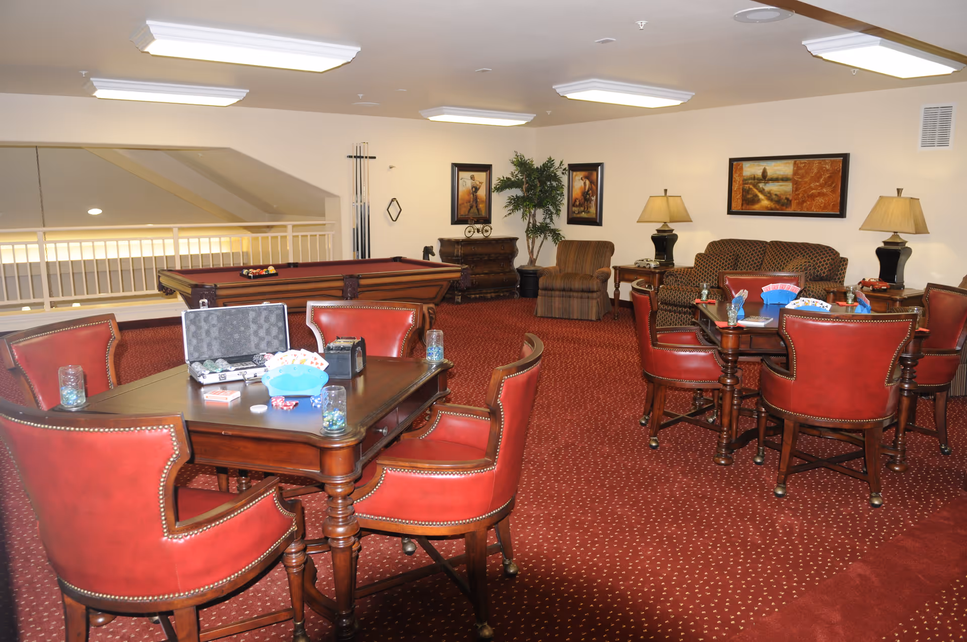 A cozy game room with two wooden tables surrounded by red leather chairs. Each table has playing cards and poker chips on it. In the background, there is a pool table with a rack of pool cues against the wall. The room has red carpet with a dotted pattern, beige walls adorned with framed artwork, and a couple of table lamps providing warm lighting. A patterned sofa and an armchair are also visible, along with a potted plant in the corner.