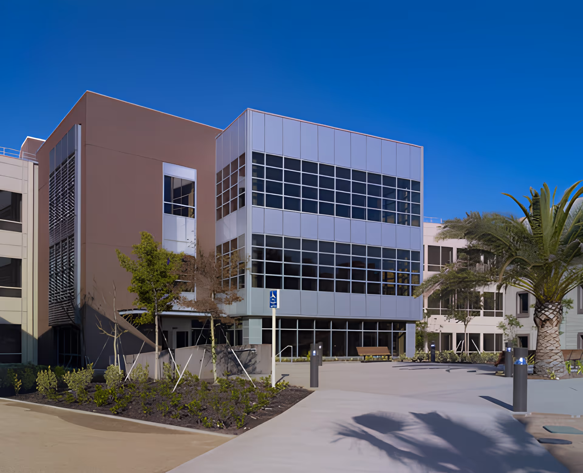 Exterior view of a modern multi-story building with large glass windows and a beige facade, surrounded by landscaped greenery including small trees and bushes, under a clear blue sky.