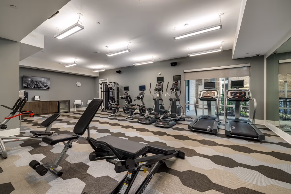 A modern fitness room with various exercise equipment including treadmills, stationary bikes, elliptical machines, and weight benches. The room has a patterned carpet floor, gray walls, bright overhead lighting, a wall-mounted TV, and large windows letting in natural light.
