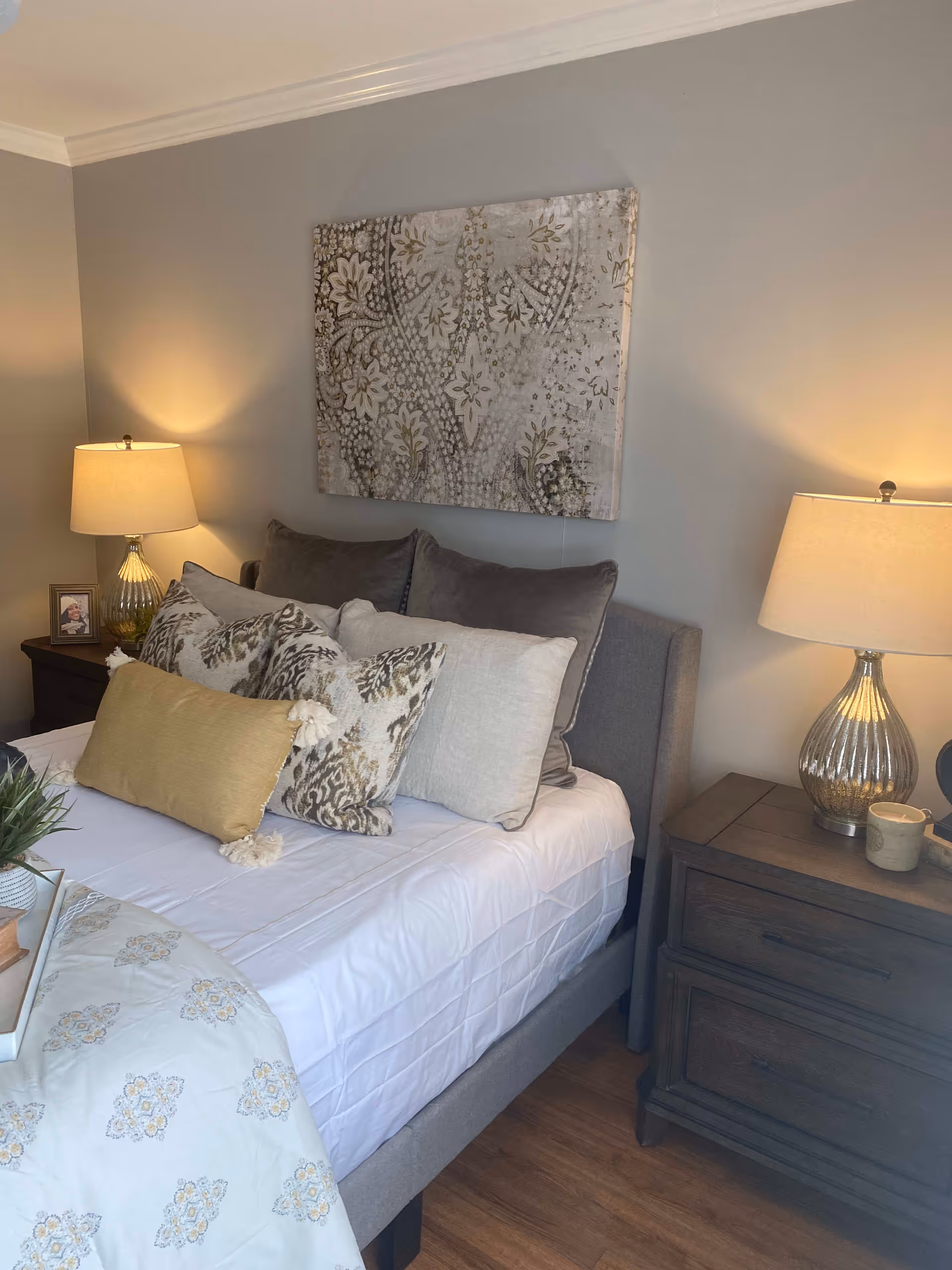 A cozy bedroom with a neatly made bed featuring multiple decorative pillows in shades of gray, beige, and yellow. There are two wooden nightstands on either side of the bed, each with a lamp emitting warm light. Above the bed hangs a large piece of artwork with a floral and paisley pattern in neutral tones. The room has light gray walls and wooden flooring.