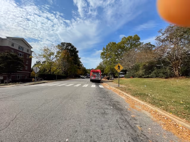A street view showing a red and white bus stopped near a pedestrian crossing sign. On the left side, there is a multi-story building partially visible with trees around it. On the right side, there is a grassy area with more trees under a partly cloudy sky. A finger is partially visible in the top right corner of the image.